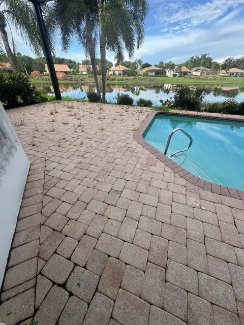 Paver pool deck overgrown with weeds, with a pool and lake beyond