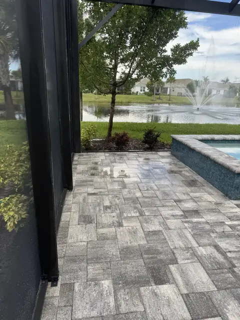 Paver patio beside a pool with a fountain beyond, seen through a screen enclosure